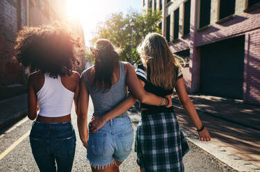 Rear view shot of three young women walking together on city street. Multiracial female friends out on the street on a summer day. - JLPSF02080
