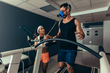 Woman giving instruction to male runner on treadmill in laboratory. Runner with mask on treadmill in laboratory with woman trainer. - JLPSF02091