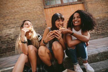 Bild von Freundinnen, die im Freien sitzen und Pizza essen. Drei junge Frauen sitzen auf dem Bürgersteig und essen Pizza. - JLPSF02093