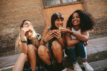 Picture of female friends sitting outdoors and eating pizza. Three young women sitting on the sidewalk and having pizza. - JLPSF02093