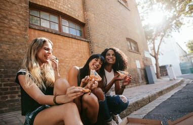 Shot of three beautiful girls sitting outdoors by the road eating pizza. Female friends relaxing by the street and smiling. - JLPSF02095