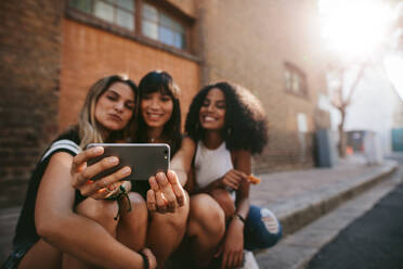 Drei lächelnde Freundinnen machen ein Selfie mit einem Smartphone. Eine multiethnische Gruppe von Frauen sitzt im Freien auf der Straße und macht ein Selbstporträt mit einem Mobiltelefon. - JLPSF02097