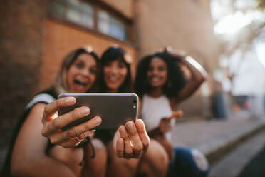 Drei Freundinnen machen ein Selfie mit dem Handy. Eine Gruppe von Frauen sitzt im Freien und macht ein Selbstporträt mit dem Smartphone. Fokus auf das Handy. - JLPSF02098