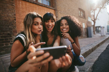 Weibliche Freunde sitzen im Freien und machen ein Selfie mit dem Smartphone. Multiethnische Gruppe von Frauen hängen in der Stadt ab und machen ein Selbstporträt mit dem Handy. - JLPSF02099