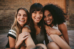 Drei schöne lächelnde Freundinnen nehmen Selfie mit Handy. Multi ethnischen Gruppe von Frauen sitzen im Freien durch die Stadt Straße und nehmen Selbstporträt. - JLPSF02100