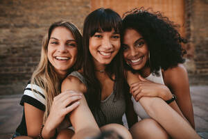 Three beautiful smiling girlfriends taking selfie with mobile phone. Multi ethnic group of women sitting outdoors by the city street and taking self portrait. - JLPSF02100