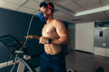 Professional athlete monitoring his performance in sports science lab. Muscular man with face mask running on treadmill in gym. - JLPSF02106