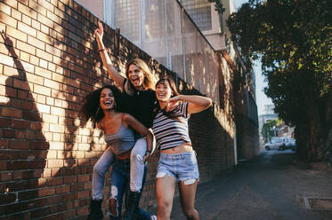 Shot of young woman carrying her female friend on her back. Three young women having fun on city street. - JLPSF02132