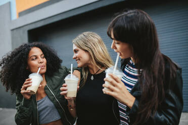 Aufnahme von drei jungen Frauen, die auf einer Straße in der Stadt Eiskaffee trinken. Verschiedene Gruppen von Freundinnen trinken Eiskaffee im Freien. - JLPSF02143