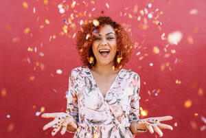 Junge Frau wirft sternförmige bunte Konfetti in die Luft. Nahaufnahme von Frau in glücklicher Stimmung mit Konfetti rundherum vor einem roten Hintergrund. - JLPSF02160