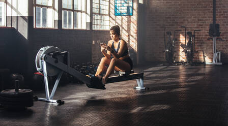 Young woman using phone while training at the gym. Woman sitting on exercising machine holding mobile phone. - JLPSF02167