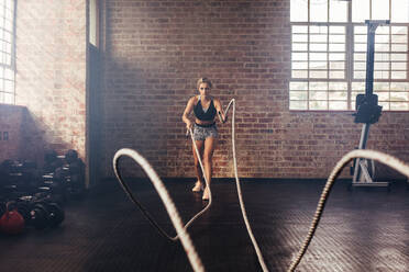 Junge Frau beim Krafttraining mit schweren Seilen im Fitnessstudio. Die Athletin bewegt die Seile in einer Wellenbewegung als Teil des Krafttrainings. - JLPSF02169