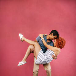 Young man carrying his girlfriend in his arms and touching his forehead with hers. Young couple in a romantic pose against red background. - JLPSF02174