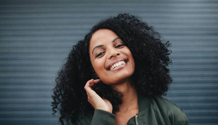 Close up Porträt der schönen afrikanischen Frau, die vor einem grauen Hintergrund steht. Afroamerikanische Frau schaut in die Kamera und lächelt. - JLPSF02191