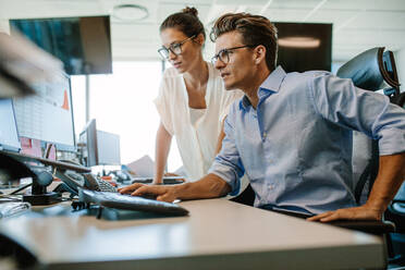 Aufnahme von zwei Geschäftskollegen, die in ihrem Büro an einem Desktop-Computer arbeiten. Der Mann sitzt an seinem Schreibtisch, seine Kollegin steht daneben. - JLPSF02202