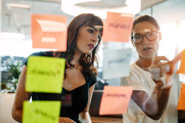 Asian businesswoman showing her coworker an idea posted on a sticky note wall. Creative professionals brainstorming on new business ideas. - JLPSF02207