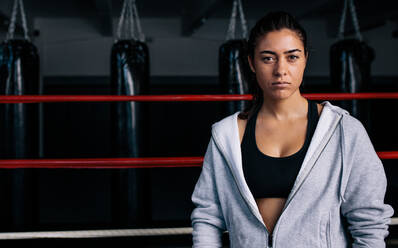 Ernst dreinblickende weibliche Boxerin in einem Boxring, die ein Sweatshirt trägt. Boxer trainiert in einem Boxstudio. - JLPSF02214