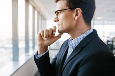 Seitenansicht eines reifen Geschäftsmannes, der an einem Bürofenster steht, nach draußen schaut und mit der Hand am Kinn nachdenkt. - JLPSF02236