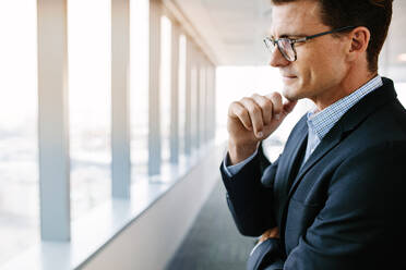 Side view shot of mature businessman standing in office and thinking with hand on chin. Caucasian male entrepreneur in suit and eyeglasses looking pensive. - JLPSF02237