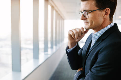 Seitenansicht eines reifen Geschäftsmannes, der im Büro steht und mit der Hand am Kinn nachdenkt. Kaukasischer männlicher Unternehmer im Anzug und mit Brille, der nachdenklich aussieht. - JLPSF02237