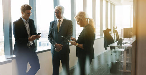 Three business people standing together in office with mobile phone and digital tablet. Corporate professional having informal meeting in modern office. - JLPSF02252