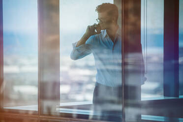 Mature business man standing inside office building and using cell phone. Man standing by window and talking on mobile phone. - JLPSF02275