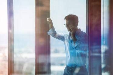 Happy businessman standing by window and talking on cell phone. Male executive inside office building and using mobile phone. - JLPSF02276