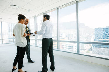 Business people standing at new office space discussing with estate broker. Real estate agent talking with potential clients inside an empty office space. - JLPSF02289