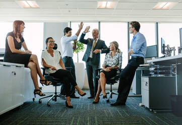 Zwei fröhliche Geschäftsleute geben sich die Hand, während ihre Kollegen sie anstarren und lächeln. Geschäftsteam genießt Erfolg im Büro - JLPSF02291
