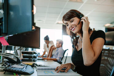 Lächelnde reife Geschäftsfrau mit Headset, die an ihrem Schreibtisch arbeitet, mit Kollegen im Hintergrund. - JLPSF02296