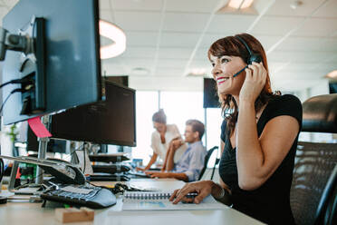 Lässige Geschäftsfrau bei der Arbeit am Schreibtisch mit Computer und Headset im Büro. Fokus auf die Frau, die im Vordergrund sitzt, während ihre Kollegen im Hintergrund arbeiten. - JLPSF02297