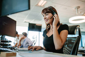 Schöne reife Geschäftsfrau, die über ein Headset spricht, während sie an ihrem Schreibtisch arbeitet. Kaukasische weibliche Führungskraft mit arbeitenden Menschen im Hintergrund. - JLPSF02298