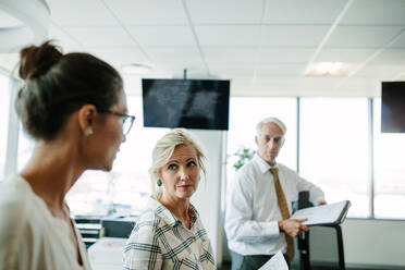 Reife Geschäftsfrau hört ihrem Kollegen zu, während ein Mann im Hintergrund steht. Geschäftsleute haben ein informelles Treffen in einem modernen Büro. - JLPSF02307