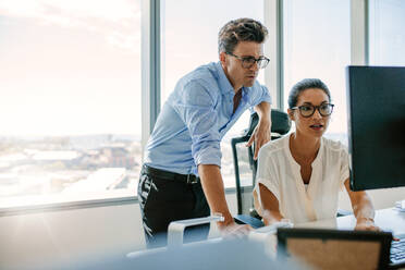 Asiatische Geschäftsfrau sitzt an ihrem Schreibtisch und arbeitet am Computer mit einem männlichen Kollegen, der daneben steht. Zwei Geschäftsleute arbeiten zusammen im Büro. - JLPSF02313