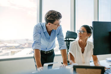 Asian businesswoman sitting at her desk and looking at male colleague standing by. Two corporate professionals working together in office. - JLPSF02314