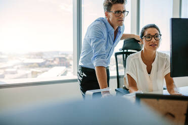 Reife Geschäftsfrau, die an ihrem Schreibtisch sitzt und mit einem männlichen Kollegen am Computer arbeitet. Zwei Geschäftsleute, die auf einen Computerbildschirm im Büro schauen. - JLPSF02316