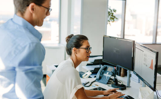 Reife Geschäftsfrau arbeitet am Computer mit einem männlichen Kollegen, der daneben steht. Geschäftsmann und Frau am Schreibtisch, die auf den Computerbildschirm schauen. - JLPSF02318