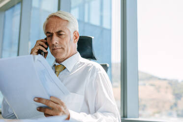Shot of mature businessman sitting at his desk reading a document and talking on mobile phone. Senior male entrepreneur working at his office desk. - JLPSF02324