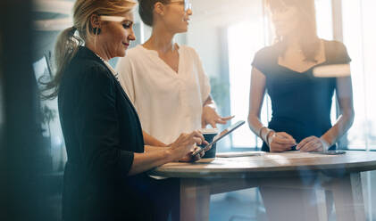 Drei weibliche Kollegen, die eine Besprechung im Stehen an einem Tisch abhalten. Ältere Frau, die ein digitales Tablet benutzt, während ihre Kollegen im Stehen diskutieren. - JLPSF02327