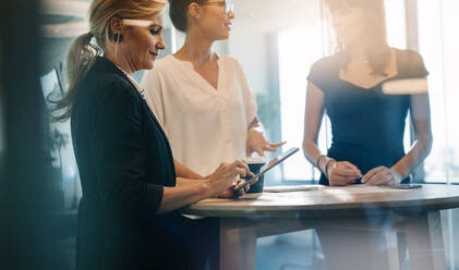 Three female colleagues having a standing meeting around a table. Mature woman using digital tablet with coworkers discussing standing by. - JLPSF02327