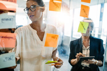 Portrait of creative professionals looking over a post it note wall and discussing. Female executives standing at the office behind glass wall with sticky notes. - JLPSF02342