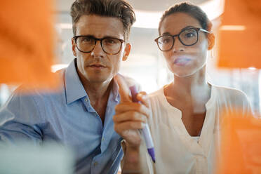 Mature businesswoman pointing at adhesive note and discussing with male colleague. Professionals brainstorming on new business ideas behind glass wall. - JLPSF02348