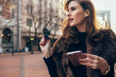 Schöne junge Frau, die mit Handy und Sonnenbrille die Straße entlanggeht. Stilvolle kaukasische Frau in der Stadt, die wegschaut. - JLPSF02379