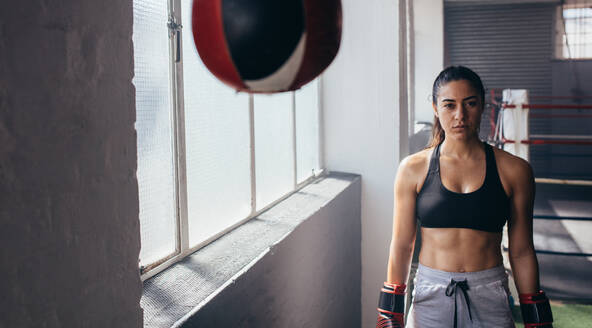 Weibliche Boxerin in einem Boxstudio, die in ihren Boxhandschuhen in der Nähe eines Boxsacks steht. - JLPSF02413