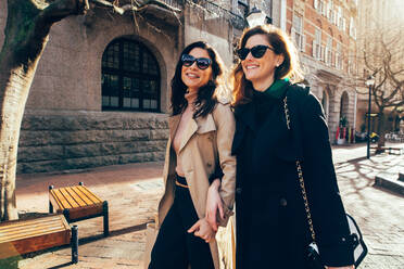 Zwei Frauen, die lächelnd im Freien spazieren gehen, Freundinnen, die zusammen auf einer Straße in der Stadt spazieren gehen. - JLPSF02414