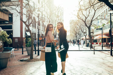 Two attractive young woman holding shopping bags walking on the street looking back and smiling. Shopaholics walk around the city. - JLPSF02423