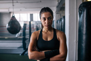 Nahaufnahme einer weiblichen Boxerin, die mit gefalteten Händen in einem Boxstudio steht. Eine Frau steht in einem Boxtrainingszentrum mit Boxsäcken und einem Boxring im Hintergrund. - JLPSF02428