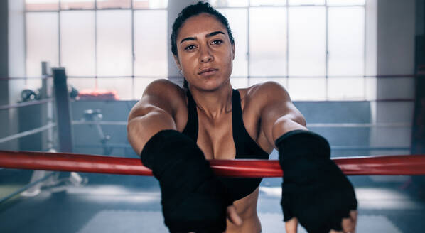 Weibliche Boxerin mit muskulösem Körper in einem Boxring stehend. Boxerin stützt ihre Arme auf das Seil des Boxrings. - JLPSF02440