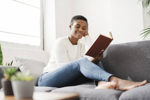 Lächelnde Frau mit geschlossenen Augen sitzt zu Hause auf der Couch und hält ein Buch - JCZF01108