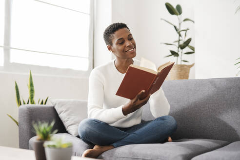 Lächelnde Frau sitzt auf der Couch zu Hause und liest ein Buch - JCZF01109